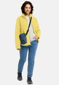 Giacca di pile gialla, t-shirt chiara, jeans blu e sneakers nere; porta una piccola borsa a tracolla blu con cerniera.