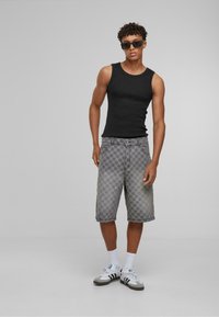 Schwarzes ärmelloses Tanktop mit grauen Shorts im hexagonalen Muster, ergänzt durch weiße Sneaker und Sonnenbrille.