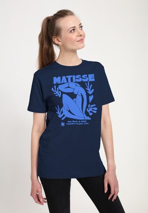 Młoda kobieta z kucykiem ubrana w granatową koszulkę Matisse z niebieską stylizowaną siedzącą nagą postacią i motywami roślinnymi.