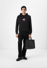 Sort hættetrøje med et logo på forsiden, sorte jeans og en sort læder tote taske. Skoene er sorte med en glat overflade. Enkel baggrund.