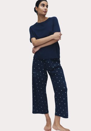 Vrouw staat blootsvoets en draagt een marineblauw shirt met korte mouwen en marineblauwe wijde broek met kleine witte patronen.