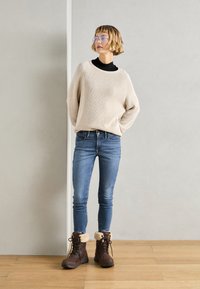 Vrouw in een beige gebreide trui, blauwe skinny jeans, bruine laarzen met bontvoering en een bril, staande binnen op een houten vloer.