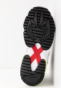 Bílé a černé podešve tenisek s texturovaným gumovým vzorem, červeným akcentem a detaily v limetkově zelené. Obsahuje černé prvky pro trakci.