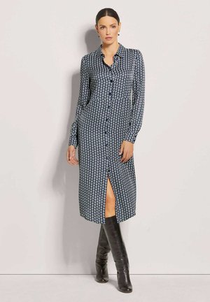 Langärmliges, marineblaues Kleid mit einem kreisförmigen weißen Muster, geknöpftem Vorderteil, gerafftem Kragen und einem hohen Schlitz bis zum Oberschenkel, kombiniert mit kniehohen schwarzen Stiefeln.