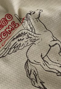 Tissu beige à texture gaufrée avec un dessin au trait noir d'un cheval ailé et un texte en gras rouge partiellement visible.