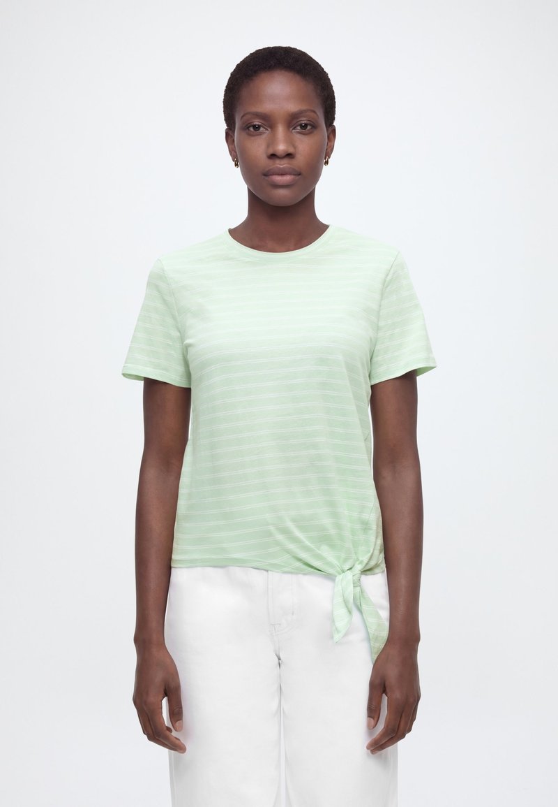 Γυναίκα που φοράει ανοιχτοπράσινο κοντομάνικο μπλουζάκι με οριζόντιες ρίγες και κόμπο στο μπροστινό μέρος, σε συνδυασμό με λευκό παντελόνι, στέκεται μπροστά σε απλό φόντο.