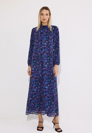 Femeie purtând o rochie cu mâneci lungi, până la gleznă, cu un model floral albastru și mov, combinată cu sandale negre cu toc înalt.