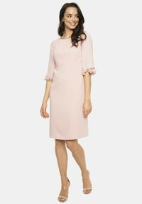 Jasnoróżowa sukienka z gładkiego materiału, z rękawami do łokcia ozdobionymi falbanami oraz prostym, do kolan krojem.