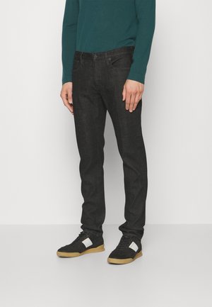 Mann trägt dunkle, eng anliegende Jeans, ein petrolfarbenes Langarmshirt und schwarze Sneaker mit weißen Details, steht vor einem schlichten weißen Hintergrund.