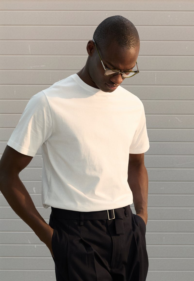 Weißes T-Shirt mit Rundhalsausschnitt aus Baumwolle, kombiniert mit dunkler Hose. Modell trägt runde Brille, steht mit den Händen in den Taschen und schaut nach unten.