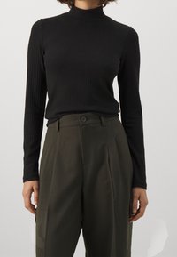 Svart ribbade långärmad topp med hög hals, åtföljd av högmidjade olivgröna byxor med knappdetalj och veckad framdel.