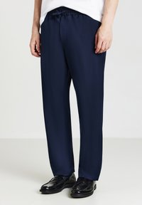 Mann in marineblauen geraden Hosen und schwarzen Lederloafers, der vor einem schlichten hellen Hintergrund steht.