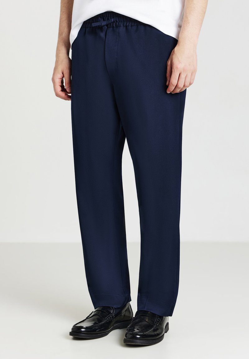 Mann in marineblauen geraden Hosen und schwarzen Lederloafers, der vor einem schlichten hellen Hintergrund steht.