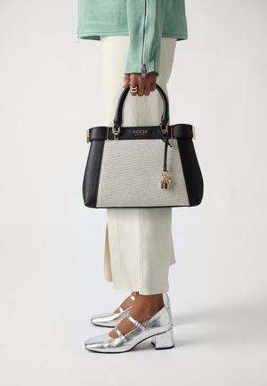 Personne tenant un sac à main Guess noir et beige avec détail de serrure dorée, portant une jupe blanche, une veste vert menthe et des chaussures argentées à talon bas.