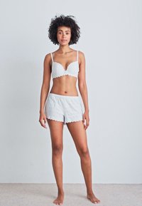 Witte kanten bralette met gegolfde randen en bijpassende shorts, met een gestructureerd patroon. Model staat voor een neutrale achtergrond.