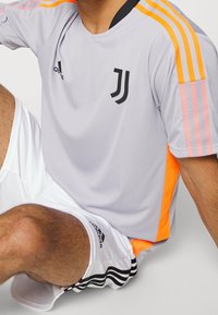 Jersey atlético gris con acentos en naranja y rosa, con un logo en el pecho y tres franjas en los hombros. Combinado con pantalones cortos blancos.