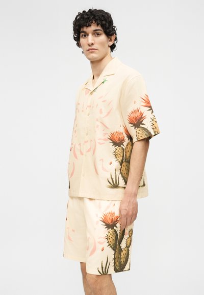 Άνδρας με σγουρά σκούρα μαλλιά φοράει μπεζ κοντομάνικο πουκάμισο με κουμπιά και αντίστοιχα σορτς με μεγάλα σχέδια κάκτου και πορτοκαλί λουλουδιού.