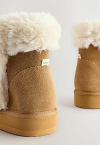 Braune Wildlederschuhe mit cremefarbener Kunstfellverzierung, flachen Gummisohlen und seitlichem Etikett. Weiche Textur und abgerundete Form.