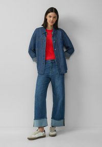 Jeansjacke mit Knopfverschluss, über einem roten strukturierten Oberteil getragen. Weite blaue Jeans mit umgeschlagenen Säumen. Sneaker mit weißen, schwarzen und tanfarbenen Akzenten.