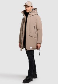 Chaleco de plumas beige con un cuello alto, bolsillos con cremallera y un dobladillo ajustable. Combinado con jeans negros y botas negras gruesas.