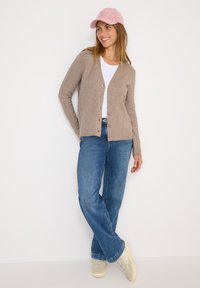 Beige kofta, vit t-shirt, blå jeans och ljusa sneakers. Koftan har v-ringning och knappar; kepsen är av rosa corduroy.