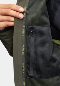 Wewnętrzna część zielonej kurtki ma siateczkową teksturę, czarną kieszeń na zamek oraz branding "ALPINUS" w żółtym kolorze wzdłuż krawędzi.