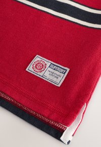 Tela roja con rayas azul marino y blancas, con un parche etiquetado que dice "Superdry Vintage Athletic Outfitters". Material de algodón, acabado texturizado.