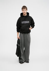 Melna kapuce ar priekšējo kabatu un grafisku tekstu, apvienota ar brīviem pelēkiem sporta biksēm un melnu nēsāšanas maisu. Gluda auduma, brīvs piegriezums.