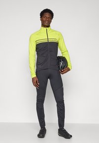 Herren-Radfahroutfit: schwarze Langarmjacke mit gelben Akzenten und reflektierenden Streifen, kombiniert mit schwarzen engen Hosen und Radschuhen.