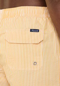 Shorts rayés jaunes et blancs avec une taille élastique, dotés d'une poche patch et d'un détail en œillets en métal. Étiquette de marque visible sur la poche.