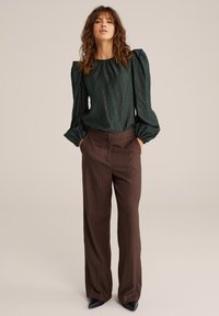 Blouse vert foncé texturé avec des manches bouffantes, associée à un pantalon marron à rayures fines taille haute et des escarpins noirs à bout pointu.