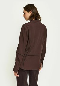 Chemise marron à manches longues en tissu texturé. Dotée d'un col classique et de coutures contrastées le long des coutures. Manches à boutons, légèrement ajustée.
