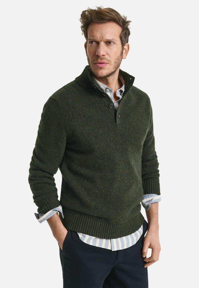 Dunkelgrüner Strickpullover mit hohem Kragen und drei Knopfverschluss. Mit gerippten Bündchen und Saum. Über einem hellblauen gestreiften Hemd getragen.