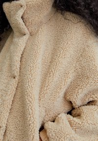 Chaqueta de forro polar beige con una superficie texturizada y suave, que cuenta con un cuello alto y pliegues en la tela para una apariencia de capas.