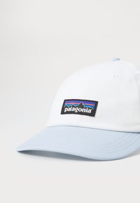 Fehér baseball sapka világoskék napernyővel, fekete téglalap alakú folttal, amely színes hegyi grafikákat és "patagonia" szöveget tartalmaz.