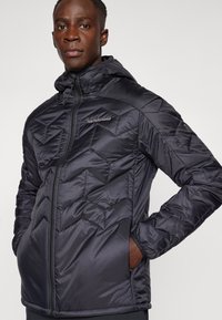 Schwarze, gestepptes Pufferjacke mit Kapuze, ausgestattet mit einem Reißverschluss vorne und Seitentaschen. Der Stoff hat eine glatte, glänzende Textur mit Zickzackmustern.