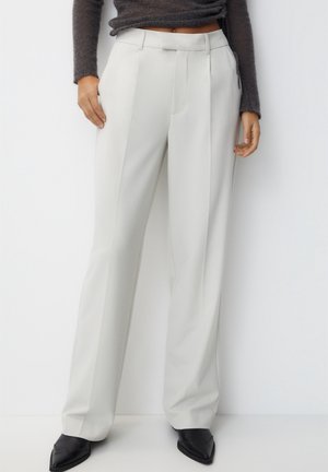 Mujer con pantalones anchos de talle alto blancos y ajustados, suéter oscuro corto y zapatos negros de punta puntiaguda sobre fondo blanco.