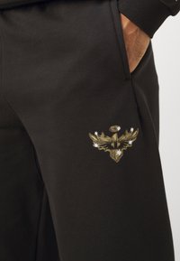 Black sweatpants featuring a textured golden embroidered emblem on the thigh, with white star accents near the design. Smooth fabric finish.