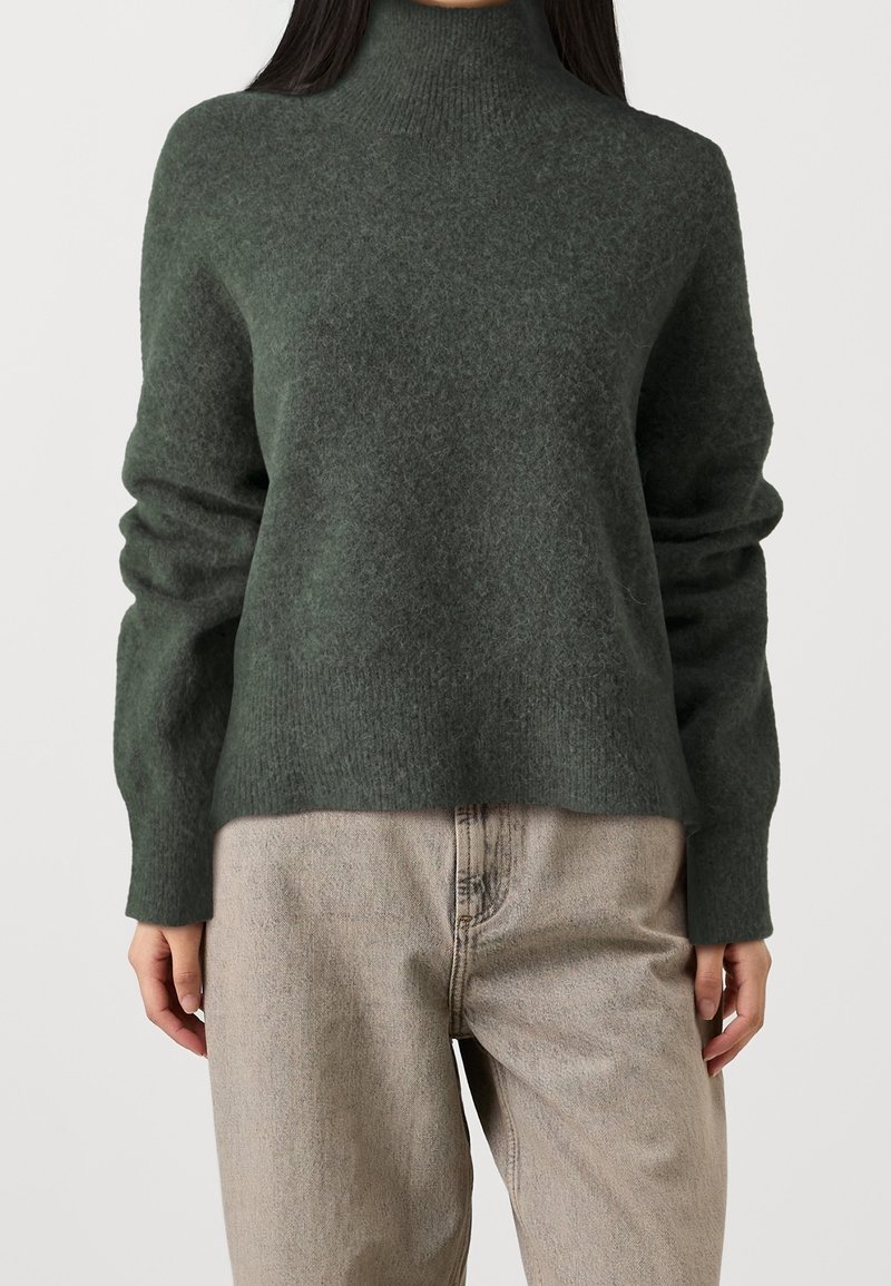 Femme portant un pull en maille douce à col roulé vert foncé et un jean ample taille haute beige clair, debout devant un fond uni.
