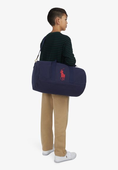 Navy blå duffel bag laget av slitesterkt stoff, med rød logo broderi, to håndtak og en justerbar skulderrem.