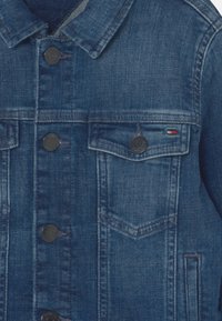 Denim jacket in dark blue with button closure, two front pockets, and collar. Features textured fabric and subtle fade detailing.