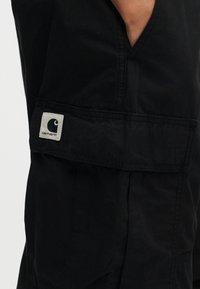 Κοντινό πλάνο στην τσέπη μαύρου παντελονιού Carhartt με ορθογώνιο καπάκι που φέρει το μπαλώμα λογότυπο Carhartt.