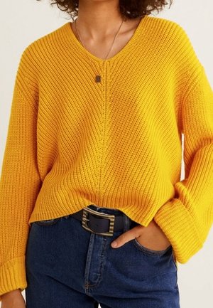 Personne portant un pull en maille jaune vif, un jean bleu foncé avec une ceinture noire, et un petit collier, avec une main dans la poche.