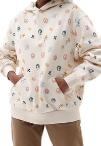 Sudadera crema con patrones coloridos que incluyen flores, signos de paz y logotipos. Cuenta con un bolsillo frontal y puños acanalados. Tejido suave.