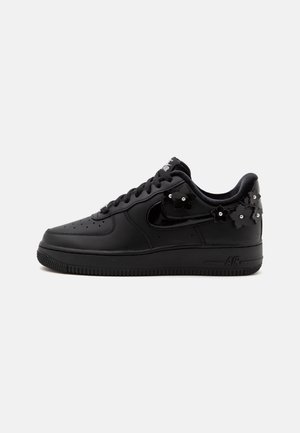 Čierny nízky teniska s šnurovaním vpredu, lesklým čiernym znakom Swoosh a malými čiernymi kvetinovými ozdobami so striebornými cvokmi po boku a na päte.