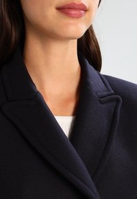 Marinblå ullrock med notched krage, texturerat tyg och knappstängning; bärs över en ljusfärgad skjorta.