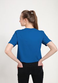 Camiseta de manga corta en algodón de color azul brillante, con un cuello redondo clásico y un corte relajado, llevada con pantalones negros, vista desde atrás.