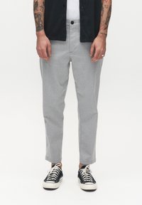 Mann in hellgrauen, cropped Hosen, schwarzen Freizeitsneakern mit weißen Schnürsenkeln, weißem Unterhemd und einem schwarzen Hemd mit Knöpfen, mit sichtbaren tätowierten Armen.