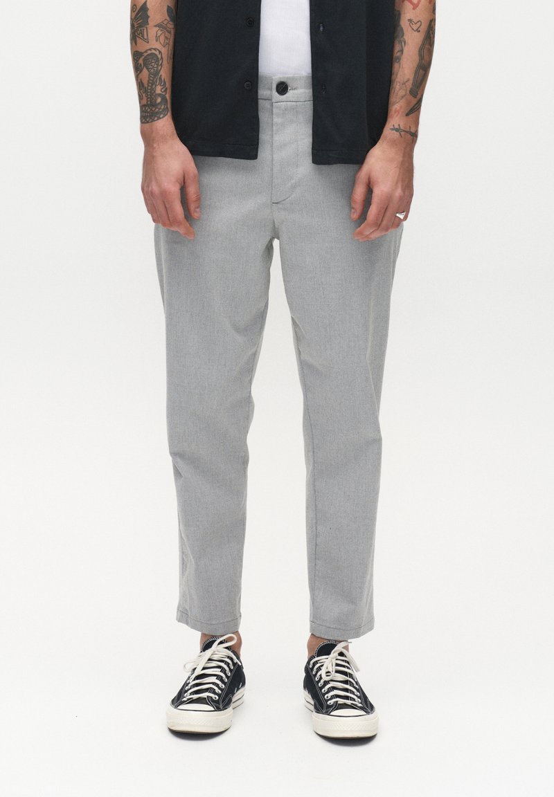 Mann in hellgrauen, cropped Hosen, schwarzen Freizeitsneakern mit weißen Schnürsenkeln, weißem Unterhemd und einem schwarzen Hemd mit Knöpfen, mit sichtbaren tätowierten Armen.