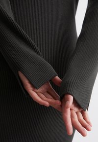 Robe noire côtelée à manches longues avec des fentes aux poignets, mettant en valeur un tissu texturé et une vue rapprochée des mains.